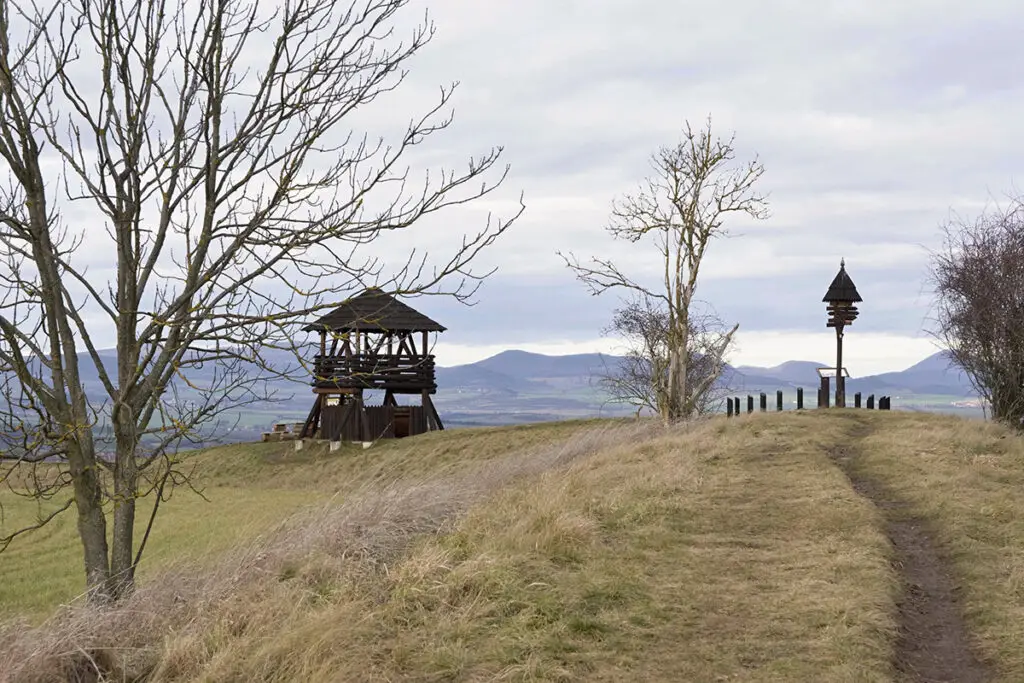 Pohled na dřevěnou rozhlednu Stradonka a dřevěný rozcestník se slunečními hodinami na valu bývalého hradiště Stradonice u Peruce. V pozadí je věnec vulkanických kup Českého středohoří pod zamračenou oblohou.