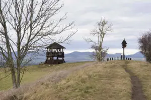 Pohled na dřevěnou rozhlednu Stradonka a dřevěný rozcestník se slunečními hodinami na valu bývalého hradiště Stradonice u Peruce. V pozadí je věnec vulkanických kup Českého středohoří pod zamračenou oblohou.