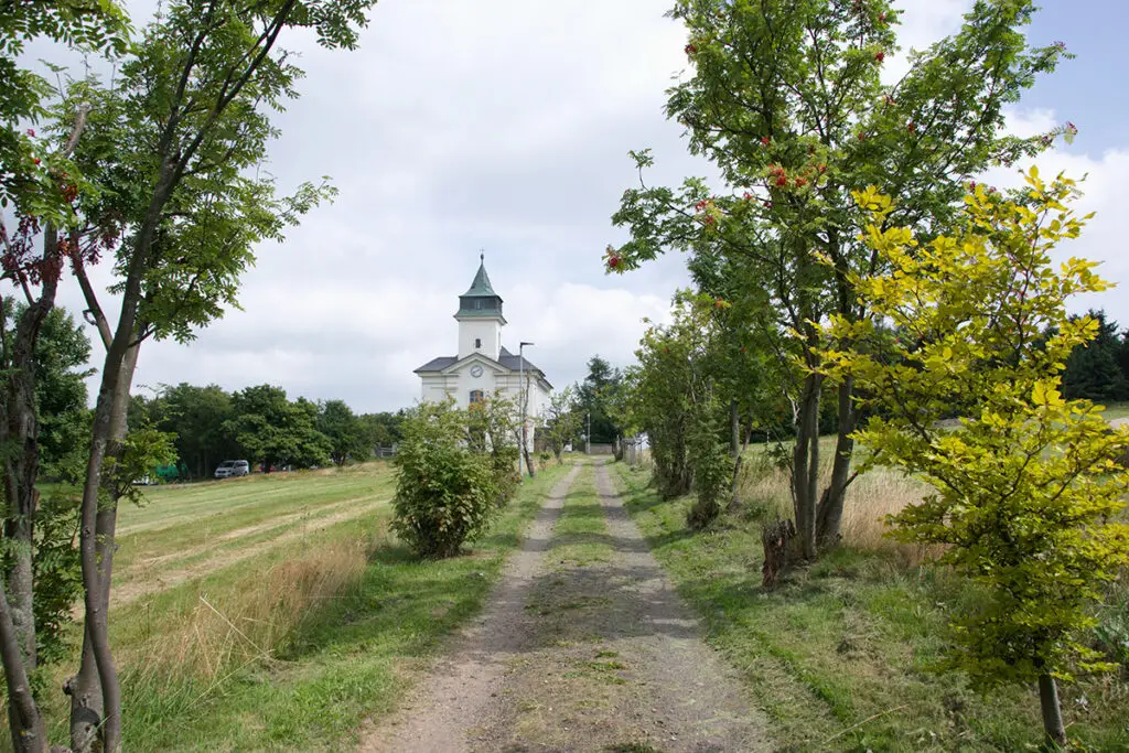 Polní cesta lemovaná stromy vedoucí k bílému kostelíku pod oblačnou oblohou – symbol cesty k vnitřnímu klidu a zastavení.