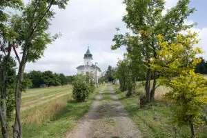 Polní cesta lemovaná stromy vedoucí k bílému kostelíku pod oblačnou oblohou – symbol cesty k vnitřnímu klidu a zastavení.