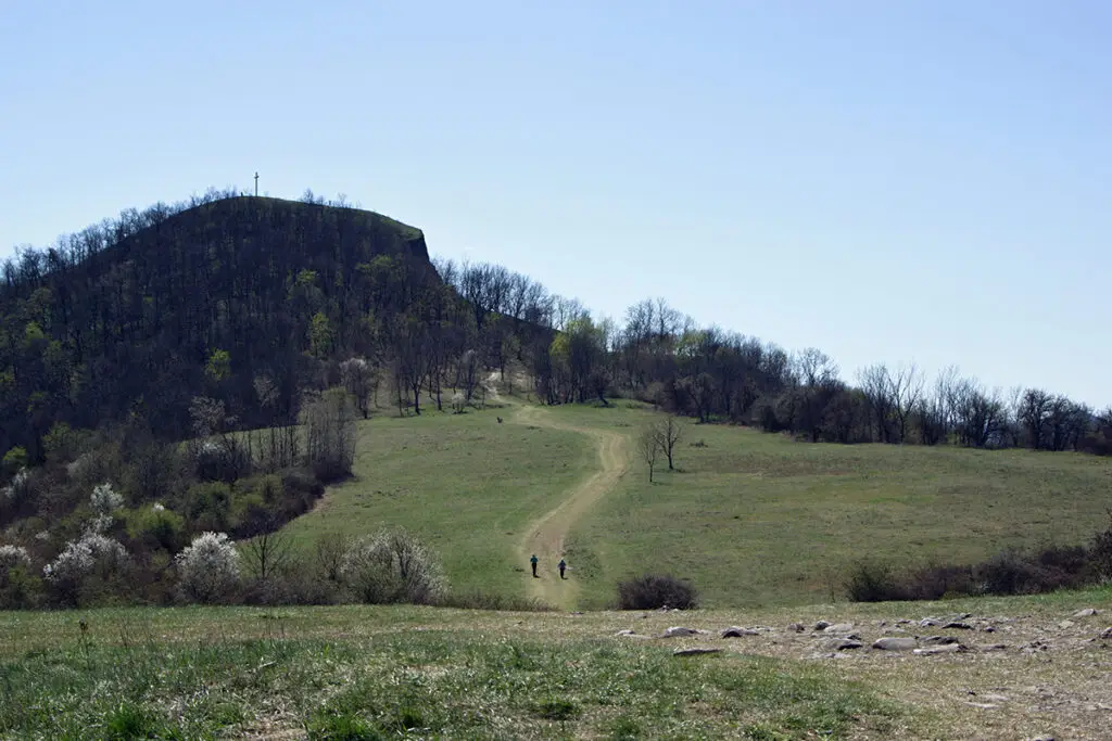 „Pohled na horu Radobýl nad Litoměřicemi s křížem na vrcholu v jarní krajině Českého středohoří. Dvě postavy jdou po polní cestě přes louku směrem k kopci pod jasnou oblohou.“