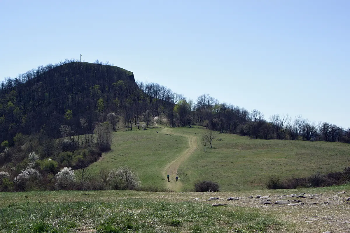 „Pohled na horu Radobýl nad Litoměřicemi s křížem na vrcholu v jarní krajině Českého středohoří. Dvě postavy jdou po polní cestě přes louku směrem k kopci pod jasnou oblohou.“