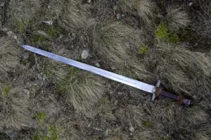 Detailní záběr na historický, kovaný středověký meč s koženou rukojetí, ležící na podzimní suché trávě a mechu, ilustrující článek o magické síle železa.