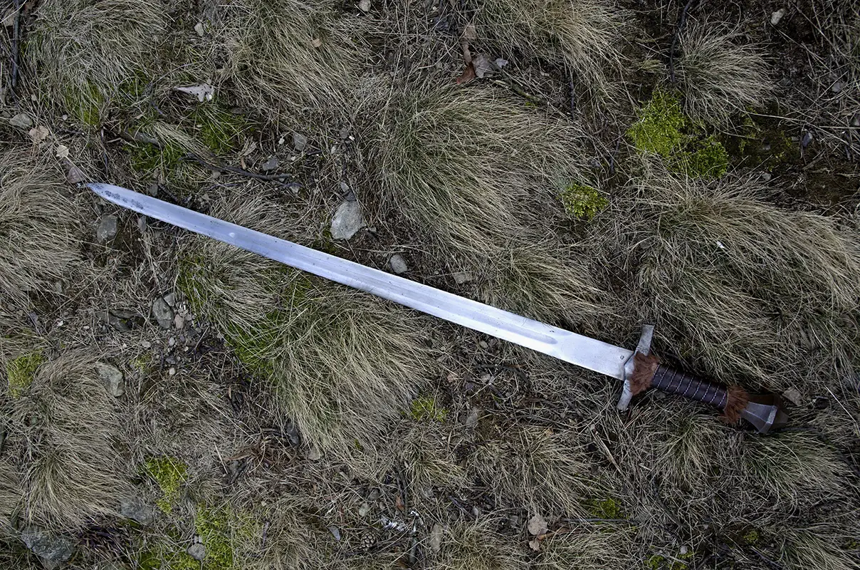 Detailní záběr na historický, kovaný středověký meč s koženou rukojetí, ležící na podzimní suché trávě a mechu, ilustrující článek o magické síle železa.
