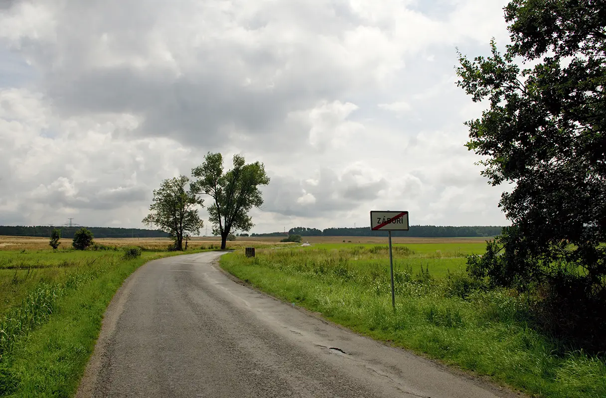 enkovská krajina v srpnu, vinoucí se silnice mezi poli a stromy, s dopravní značkou "Záboří" na kraji, pod zamračenou oblohou, zachycující atmosféru lidových pranostik o počasí.